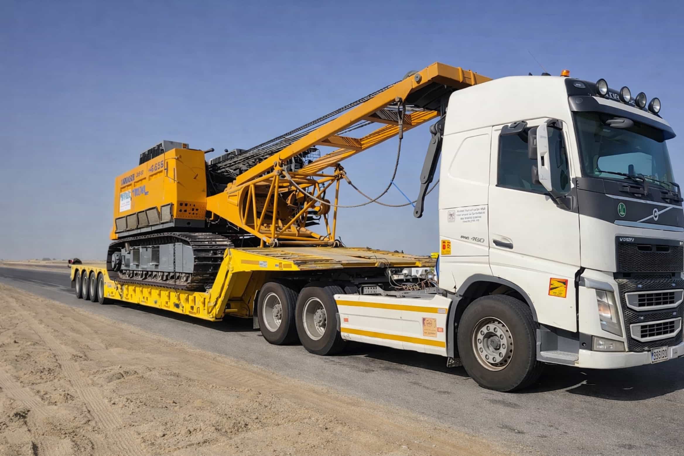 Yellow crawler crane on lowbed trailer with white Volvo truck in desert
